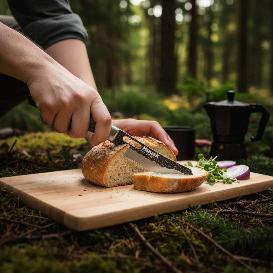 Huusk camo folding bread knife slicing a loaf on an outdoor cutting board.