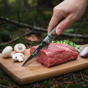 Huusk camo folding paring knife slicing raw beef at an outdoor campsite.