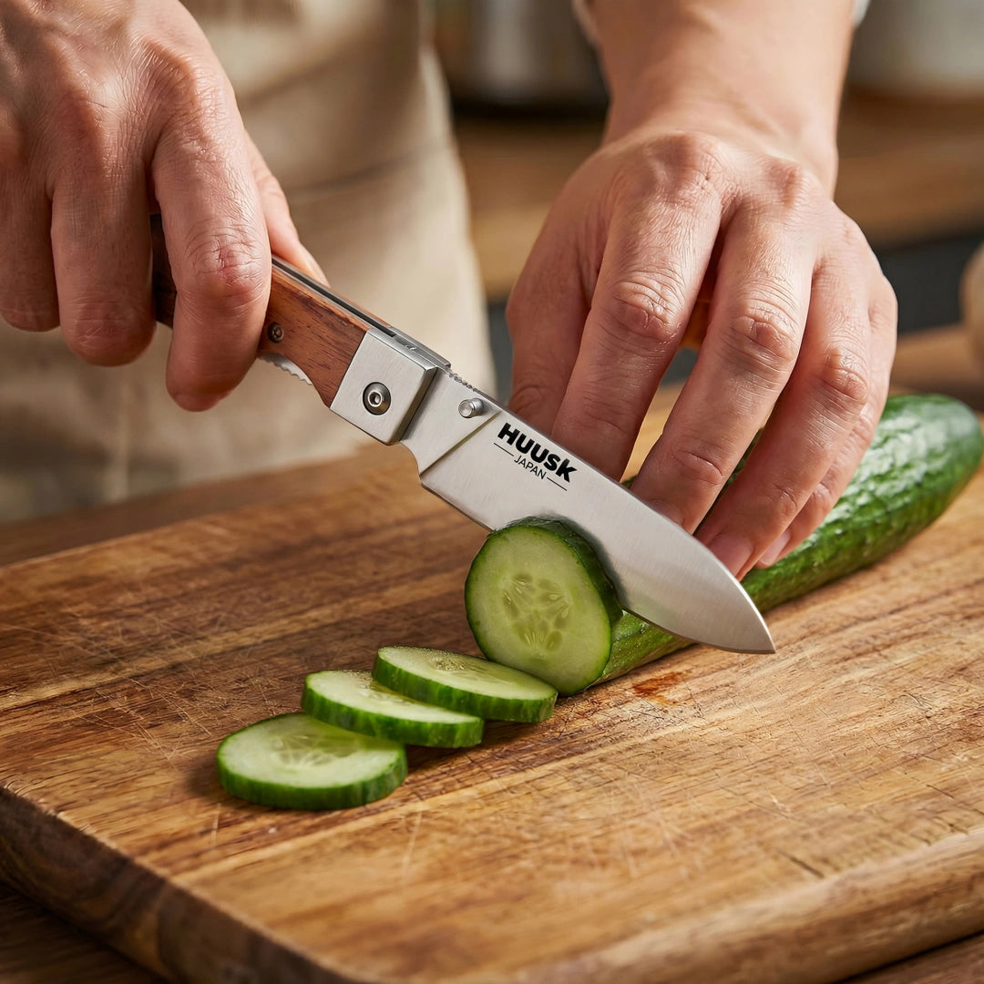 Huusk natural wood folding chef knife slicing cucumber on a kitchen cutting board.