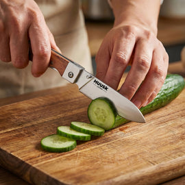 Huusk natural wood folding chef knife slicing cucumber on a kitchen cutting board.