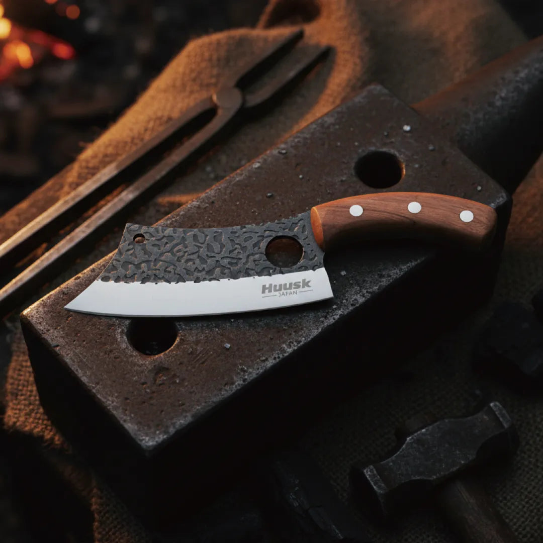 Huusk hand-forged chef knife resting on an anvil beside blacksmith tools and glowing coals.