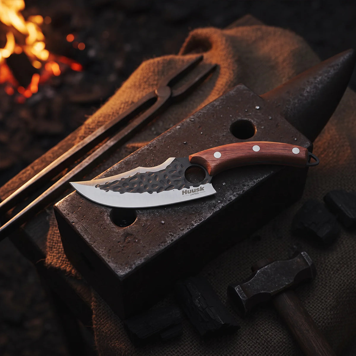 Huusk hand-forged utility knife resting on an anvil near blacksmith tools and glowing coals.