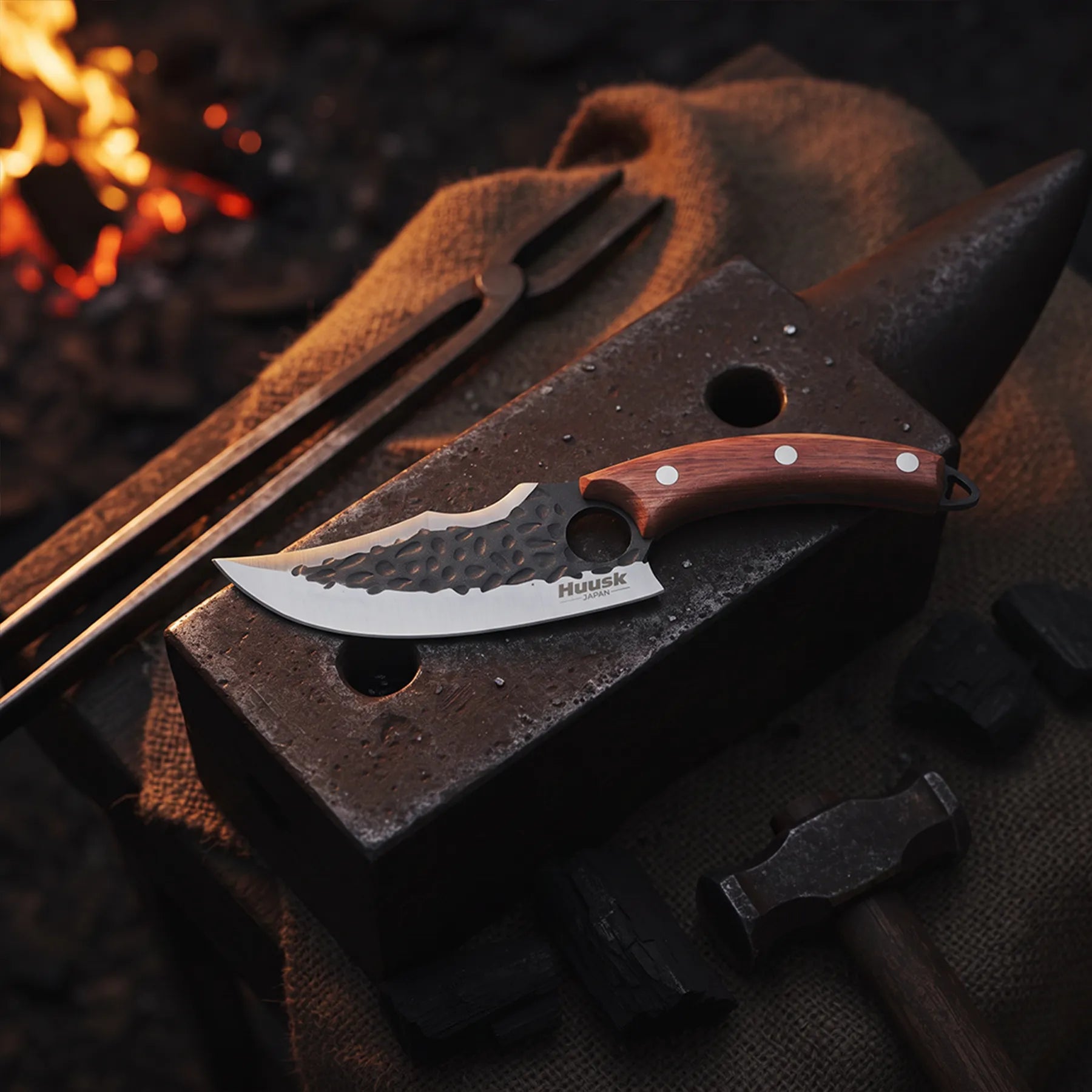 Huusk hand-forged utility knife resting on an anvil near blacksmith tools and glowing coals.